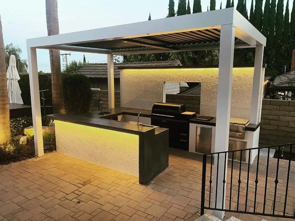 Modern illuminated outdoor kitchen beneath a white pergola with paver patio